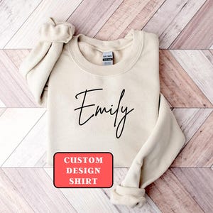 May include: A beige sweatshirt with the name "Emily" in black script. The text "CUSTOM DESIGN SHIRT" is in a red rectangle at the bottom. The sweatshirt is folded on a wooden surface.