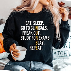 May include: Black sweatshirt with a yellow graphic that reads "EAT. SLEEP. GO TO CLINICALS. FREAK OUT. STUDY FOR EXAMS. SLAY. REPEAT."