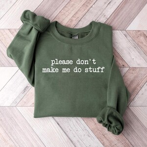 May include: Olive green sweatshirt with the phrase "please don't make me do stuff" printed in white. The sweatshirt has a crew neck and long sleeves with ribbed cuffs. The garment is laid flat on a light wood-grain surface.
