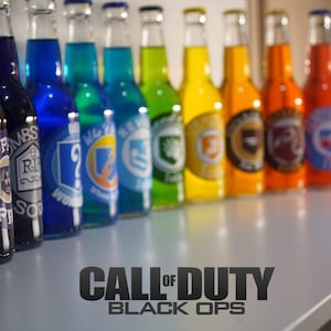 Peut inclure: Une rangée de bouteilles en verre colorées avec différentes étiquettes, dont "Call of Duty: Black Ops". Les bouteilles sont disposées sur une surface blanche.