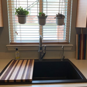 May include: A black kitchen sink with a wooden cutting board with striped wood grain. There are three hanging plants in white pots above the sink.