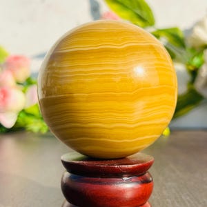 May include: A polished, spherical decorative object with a yellow and cream banded pattern, resting on a three-tiered, dark red wooden stand. The sphere's surface reflects light, and the stand has a glossy finish.