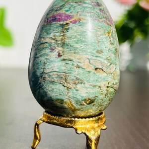 Könnte beinhalten: Ein polierter, eiförmiger Stein mit einem grün-violetten Marmormuster. Der Stein wird auf einem kleinen, verzierten, goldfarbenen Ständer präsentiert. Der Edelstein hat eine glatte, glänzende Oberfläche und ist ein dekorativer Artikel.