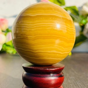May include: A polished, spherical decorative object with a banded pattern in shades of yellow and cream. The sphere rests on a dark red, wooden, three-tiered stand. The overall aesthetic is natural and elegant.
