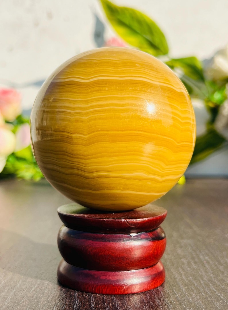 May include: A polished, spherical decorative object with a banded pattern in shades of yellow and cream. The sphere rests on a dark red, wooden, three-tiered stand. The overall aesthetic is natural and elegant.