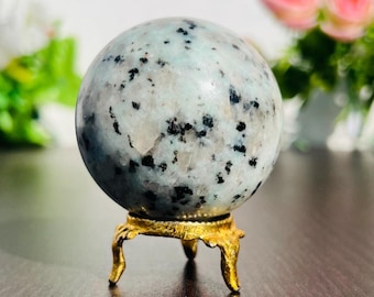 May include: A speckled, light blue and black sphere, possibly a crystal ball, sits on a small, ornate gold-colored stand. The sphere has a mottled appearance with dark spots. The stand has three curved legs and a decorative base.