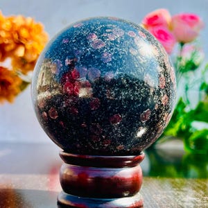 Puede incluir: Una esfera de piedra preciosa negra y roja sobre un soporte de madera. La esfera está pulida y tiene un aspecto moteado.