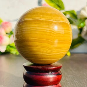 May include: A polished, golden-yellow sphere with horizontal banding rests on a three-tiered, dark red wooden stand. The sphere's surface reflects light, and the stand has a glossy finish. The background includes blurred greenery and flowers.