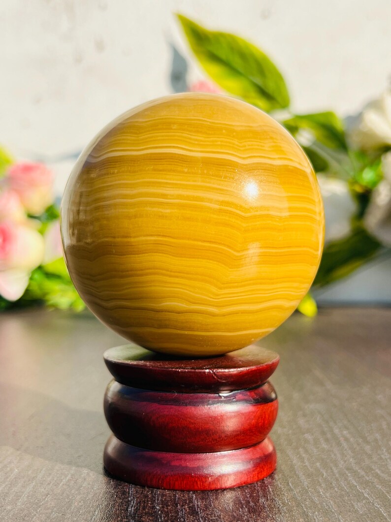 May include: A polished, golden-yellow sphere with horizontal banding rests on a three-tiered, dark red wooden stand. The sphere's surface reflects light, and the stand has a glossy finish. The background includes blurred greenery and flowers.