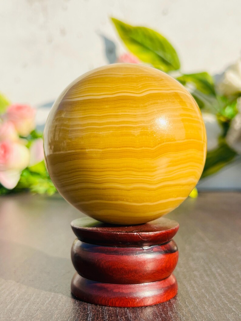 May include: A polished, spherical decorative object with a yellow and cream banded pattern, resting on a three-tiered, dark red wooden stand. The sphere's surface reflects light, and the stand has a glossy finish.