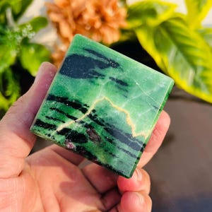 Puede incluir: Una piedra preciosa pulida de color verde, negro y blanco con forma cuadrada. La piedra preciosa tiene una superficie lisa y pulida y es un cristal natural.
