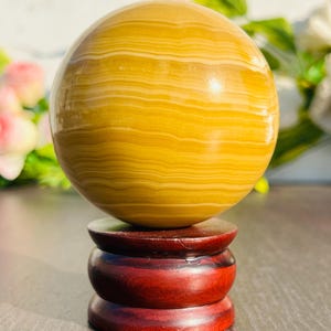 May include: A golden-yellow sphere with horizontal banding, displayed on a three-tiered, dark red wooden stand. The sphere has a polished, glossy finish. The stand is made of polished wood.
