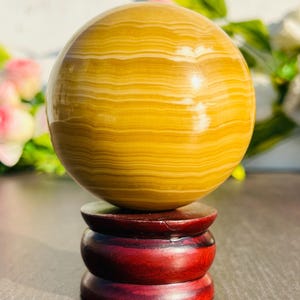 May include: A polished, golden-yellow sphere with horizontal banding, displayed on a three-tiered, dark red wooden stand. The sphere has a smooth, reflective surface. The stand is a deep, rich red color.