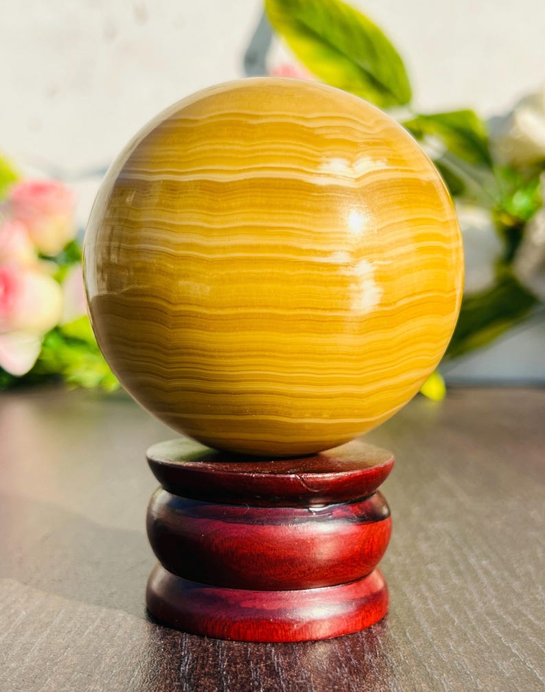 May include: A polished, golden-yellow sphere with horizontal banding, displayed on a three-tiered, dark red wooden stand. The sphere has a smooth, reflective surface. The stand is a deep, rich red color.