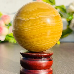 May include: A polished, spherical gemstone in shades of yellow and cream, resting on a dark red wooden stand. The orb's surface displays natural banding patterns. The stand has a tiered design, adding to the decorative appeal of the crystal ball.