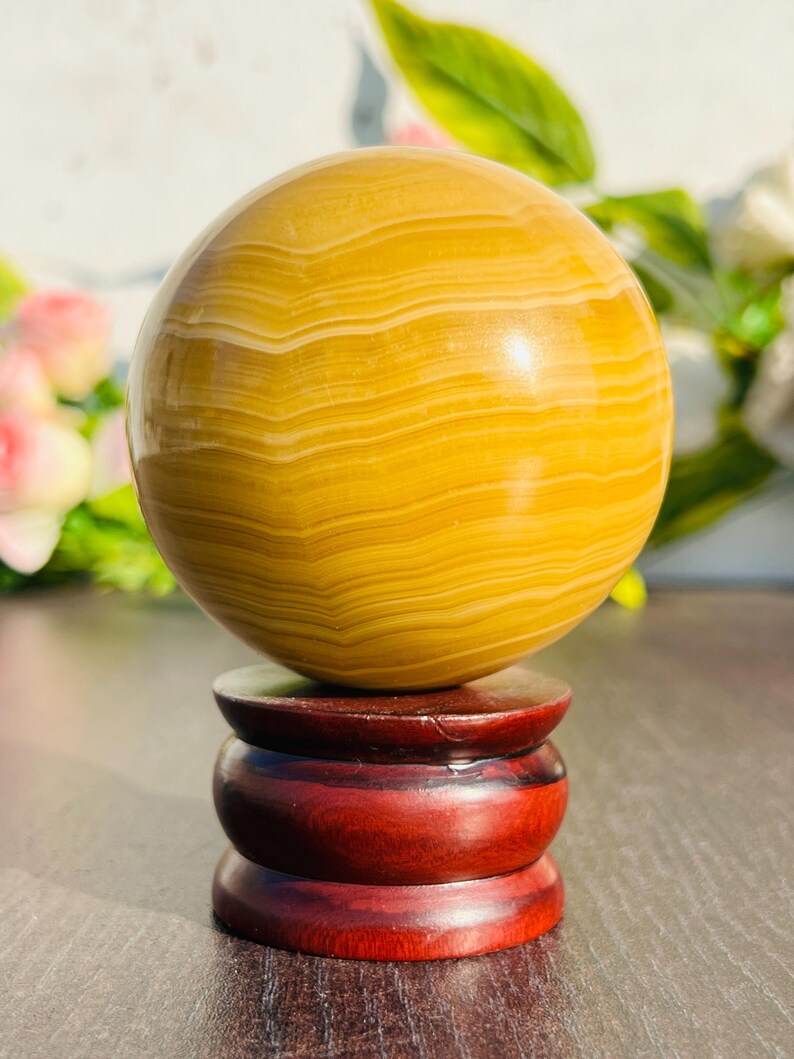 May include: A polished, spherical gemstone in shades of yellow and cream, resting on a dark red wooden stand. The orb's surface displays natural banding patterns. The stand has a tiered design, adding to the decorative appeal of the crystal ball.