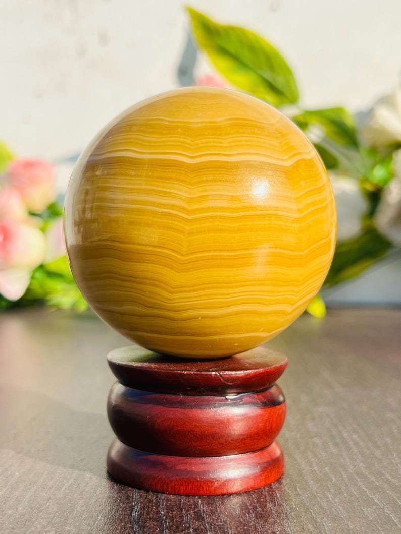 May include: A polished, golden-yellow sphere with banded patterns, displayed on a tiered, dark red wooden stand. The sphere's surface reflects light, showcasing its natural variations. The stand provides a stable base for this decorative object.