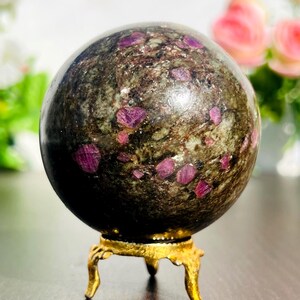 May include: A polished gemstone sphere with a dark green and brown matrix, speckled with vibrant pink ruby crystals. The sphere rests on a small, ornate gold-colored stand with three legs. A decorative item.