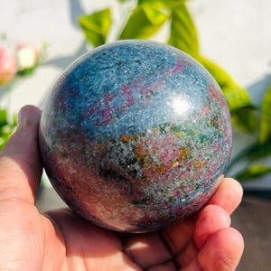 May include: A polished gemstone sphere, held in a hand, displays a mix of colors. The sphere's surface features a blend of blue, pink, and green hues, creating a marbled effect. The gemstone is a decorative item.