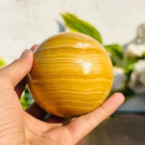 May include: A golden-yellow sphere with horizontal banding, held in a hand. The polished stone has a smooth surface and a warm color palette, with light and dark yellow tones. The sphere is a decorative item.