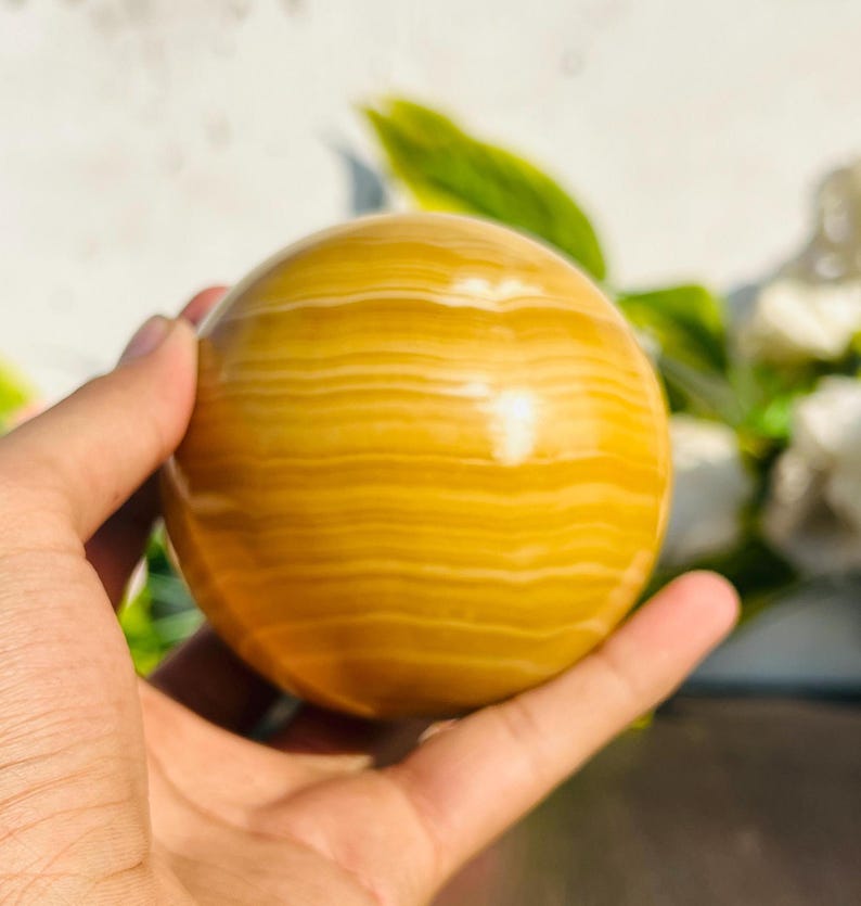 May include: A golden-yellow sphere with horizontal banding, held in a hand. The polished stone has a smooth surface and a warm color palette, with light and dark yellow tones. The sphere is a decorative item.
