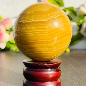 May include: A polished, spherical gemstone in shades of yellow and cream, resting on a three-tiered, dark red wooden stand. The orb's surface displays a banded pattern. The stand has a glossy finish, enhancing the overall presentation.
