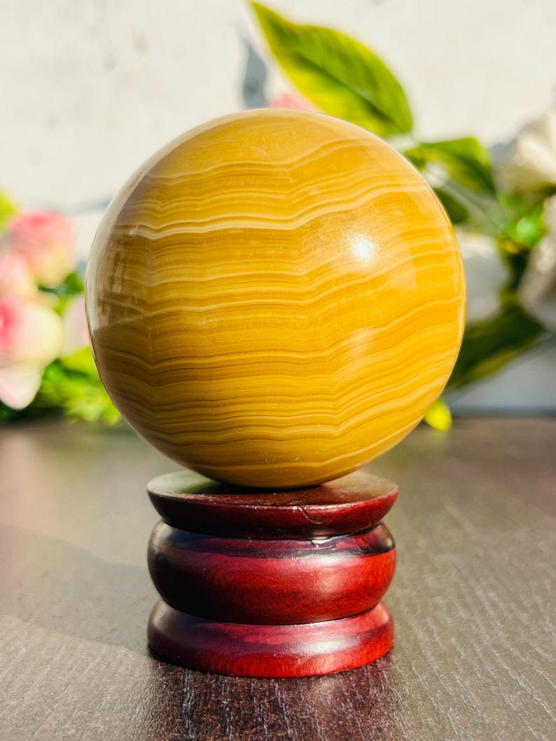 May include: A polished, spherical gemstone in shades of yellow and cream, resting on a three-tiered, dark red wooden stand. The orb's surface displays a banded pattern. The stand has a glossy finish, enhancing the overall presentation.
