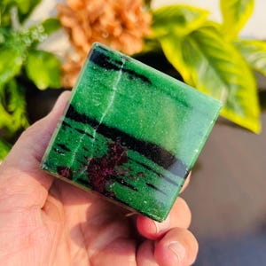 Może przedstawiać: Zielony, czarny i fioletowy sześcian z kamienia szlachetnego o gładkiej, wypolerowanej powierzchni. Sześcian ma około 5 cm.