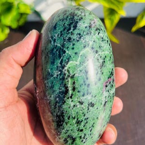 以下が含まれることがあります： 深緑、黒、赤色の斑点模様が施された、磨かれた楕円形の宝石。滑らかで丸みを帯びた形が特徴で、手で持たれています。宝石の表面はまだら模様です。