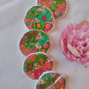 May include: A collection of five round, reusable nursing pads. Each pad features a colorful floral pattern with pink, orange, green, and white flowers. They have a white, fluffy border and a pink snap closure. A large pink flower is in the background.