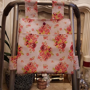 May include: A floral patterned fabric bag attached to a silver walker. The bag has a pink background with red, yellow, and green flowers. The bag has a small pink button.