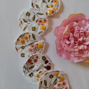 May include: Five white fabric menstrual pads with a floral print in shades of orange, yellow, and pink. The pads have a white, fuzzy trim and a yellow button on each.