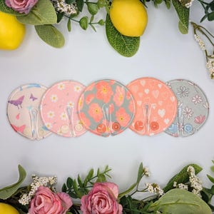 May include: Five circular fabric nursing pads with a central slit and snap closures. The patterns include butterflies on light pink, white daisies on pink, orange flowers on grey, white hearts on peach, and white flowers on grey fabric.