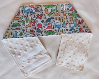 May include: White baby bib with a dinosaur print. The bib has a white, padded lining and adjustable velcro closures.