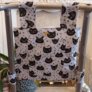 May include: A white walker with black cat faces and stars printed on it. The walker has two straps that attach to the top of the walker. The straps are made of the same fabric as the walker. The walker is in front of a white fireplace.