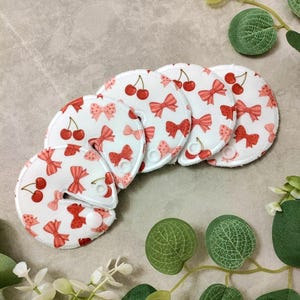May include: A set of five white, round nursing pads with a repeating pattern of red cherries and bows. Each pad has a curved edge and a snap closure. The pads are arranged on a light gray surface with green foliage.
