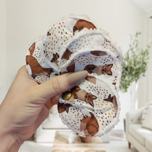 May include: A set of five white cotton reusable makeup remover pads with a scalloped edge. The pads feature a brown and gold fox print.