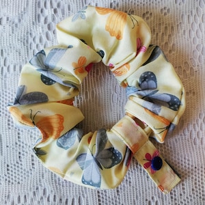 May include: A yellow scrunchie with a butterfly print and a blue button. The scrunchie has a yellow strap with a blue button.