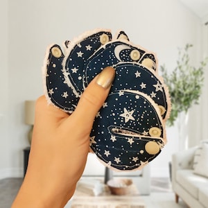 May include: Four reusable cloth menstrual pads with a dark blue background and white stars and crescent moon pattern. The pads have white snaps and a soft, white, fuzzy trim.