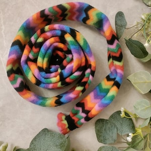 May include: A colorful, rainbow-patterned fleece toy for pets, coiled on a neutral surface. The toy features a zig-zag design in shades of purple, blue, green, orange, and red. The ends are finished with black and red accents.