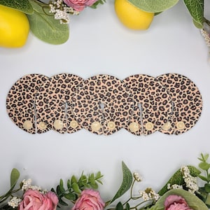 May include: Five circular fabric pads featuring a brown and black leopard print pattern. Each pad has a single slit extending from the outer edge to the centre, secured by a small, round, beige plastic snap fastener at the bottom of the slit.