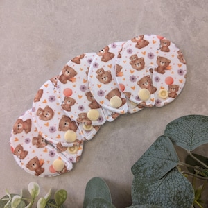 May include: A set of five circular white fabric pads featuring a repeating pattern of brown teddy bears, small purple flowers, and orange hearts. Each pad has a slit leading to the centre and a single yellow plastic snap button for closure.