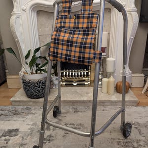 May include: A silver metal walker with four wheels and grey rubber hand grips. Attached to the front frame is a rectangular fabric pouch featuring a plaid pattern in shades of orange, navy blue, and white, secured with a single yellow snap button.