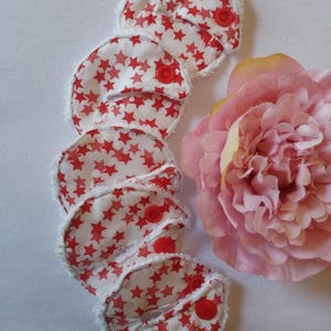 May include: Six white reusable makeup remover pads with red stars and a red button on each pad. The pads are stacked on top of each other and are surrounded by a white background.
