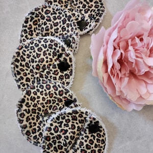 May include: A collection of round, reusable makeup remover pads with a leopard print pattern. Each pad has a black snap closure and white trim. A large pink peony flower is in the background.