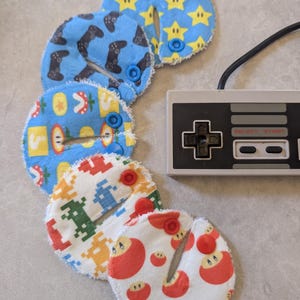 May include: A selection of patterned nursing pads featuring video game designs, such as stars, game controllers, and apples. Each pad has a button fastening. A retro games console controller is also visible.