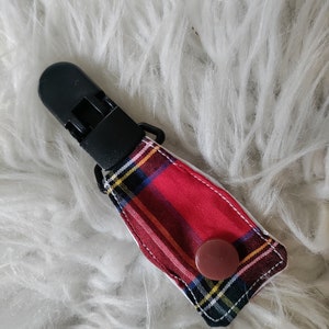 May include: A red and black plaid fabric covered clip with a black plastic clasp and a brown button.