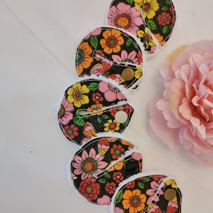 May include: Six reusable makeup remover pads with a black background and colourful floral print. The pads have a white, fluffy trim and a snap closure.