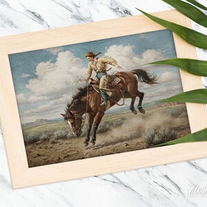 Puede incluir: Una pintura de estilo vintage de un vaquero montando un caballo marr&oacute;n. El vaquero lleva una camisa marr&oacute;n claro y pantalones vaqueros. El caballo est&aacute; dando coces y levantando polvo. La pintura est&aacute; enmarcada en un marco de madera claro.
