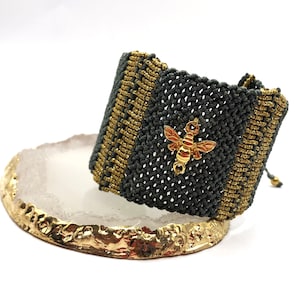 Peut inclure: Un bracelet tissé gris et or avec un charme d'abeille en or. Le bracelet est sur une tranche d'agate blanche avec un bord doré.